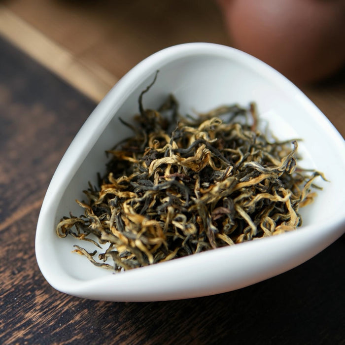 Dried loose-leaf tea leaves in a small white ceramic dish, set on a dark wooden surface.