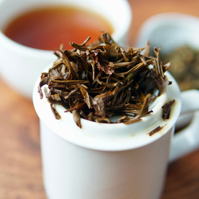 Close-up of loose-leaf tea leaves piled on the rim of a white ceramic mug, with a cup of brewed tea blurred in the background on a wooden surface.