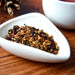 A close-up of loose-leaf herbal tea or spice blend in a small white dish, set on a wooden surface with a blurred white cup in the background.