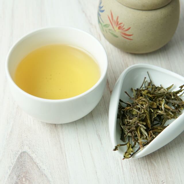 A white ceramic cup filled with pale yellow tea sits on a light wood surface beside a small white dish holding loose dried tea leaves, with part of a beige ceramic tea container visible in the background.