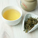 A white ceramic cup filled with pale yellow tea sits on a light wood surface beside a small white dish holding loose dried tea leaves, with part of a beige ceramic tea container visible in the background.