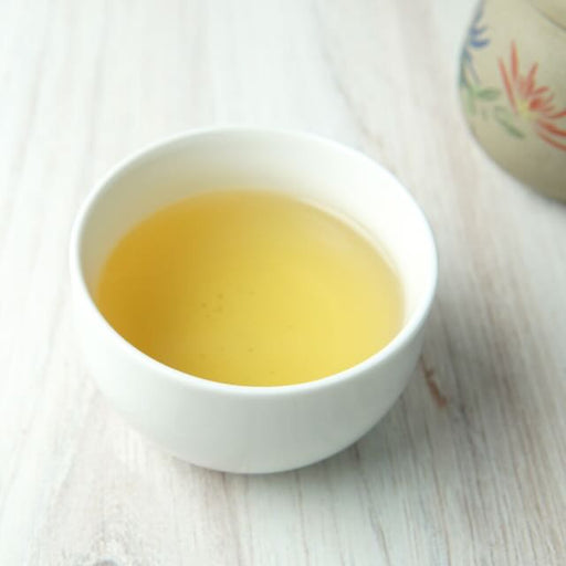 A small white ceramic cup filled with pale yellow tea, set on a light wood surface with a blurred floral ceramic object in the background.