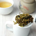 Loose-leaf green tea leaves piled on the rim of a white ceramic mug, with a cup of brewed tea and a floral ceramic container blurred in the background.