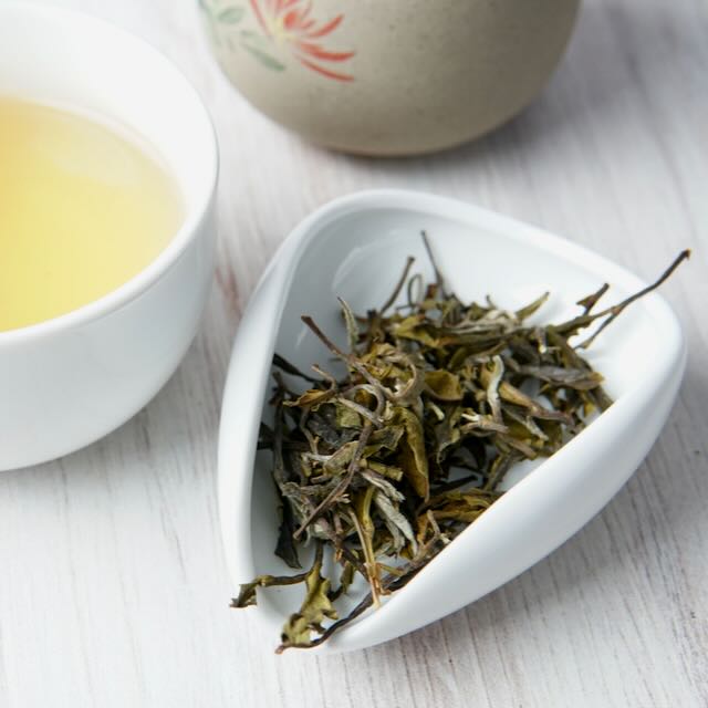 Dried loose-leaf tea leaves in a white triangular dish, with a cup of pale yellow tea and a ceramic teapot blurred in the background on a light wood surface.