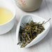 Dried loose-leaf tea leaves in a white triangular dish, with a cup of pale yellow tea and a ceramic teapot blurred in the background on a light wood surface.