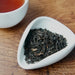 A close-up of loose-leaf tea leaves in a small white dish on a wooden surface, with part of a white cup containing amber tea visible in the upper left.