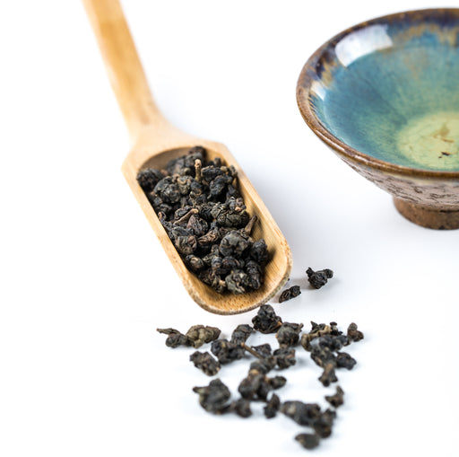 Dark roasted tea leaves scattered on a white surface beside a wooden scoop holding more tea, with a blue glazed ceramic bowl in the background.