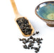 Dark roasted tea leaves scattered on a white surface beside a wooden scoop holding more tea, with a blue glazed ceramic bowl in the background.