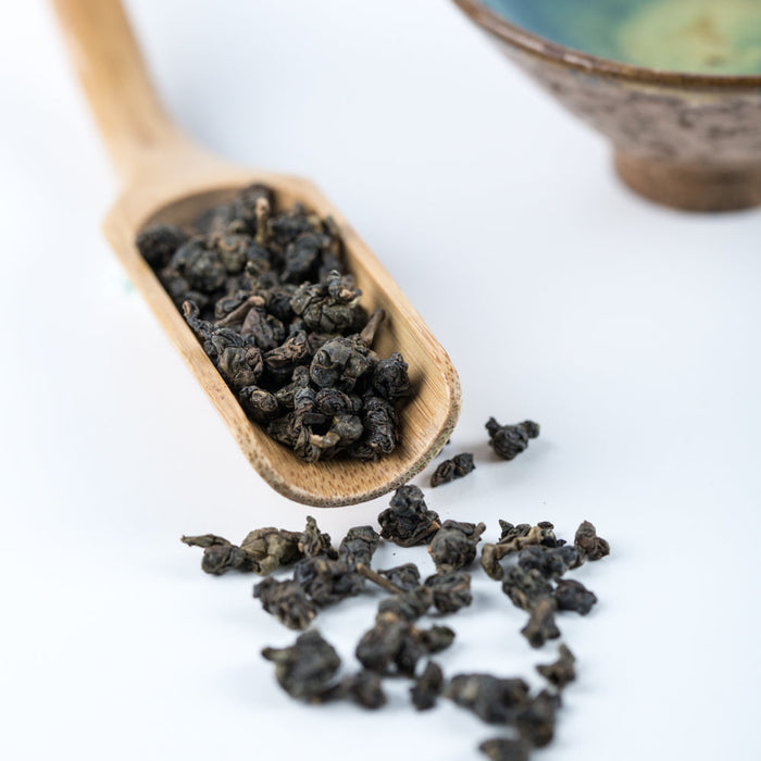 Dried dark tea leaves scattered on a white surface beside a wooden spoon holding some of the tea, with a blurred ceramic bowl in the background.