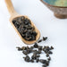 Dried dark tea leaves scattered on a white surface beside a wooden spoon holding some of the tea, with a blurred ceramic bowl in the background.