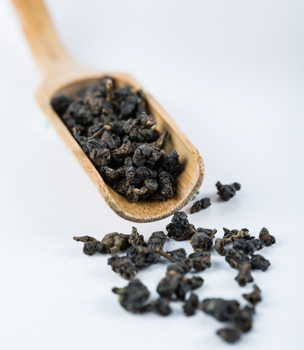 Dark roasted whole tea leaves spilling from a light wooden scoop on a clean white background.