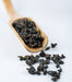 Dark roasted whole tea leaves spilling from a light wooden scoop on a clean white background.