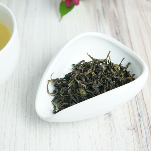 Dry green tea leaves in a white triangular dish on a light wooden surface.