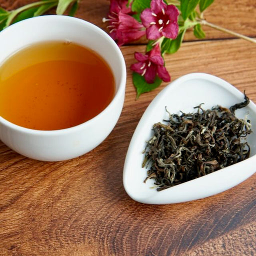 Tea cup with brewed tea and a dish of dry tea leaves on a wooden surface with flowers.