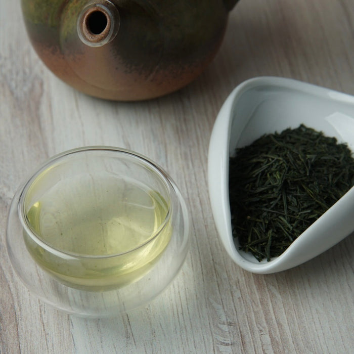 A glass cup of pale tea and a white scoop filled with loose-leaf tea on a light wood surface, with part of a ceramic teapot visible in the background.