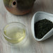 A glass cup of pale tea and a white scoop filled with loose-leaf tea on a light wood surface, with part of a ceramic teapot visible in the background.