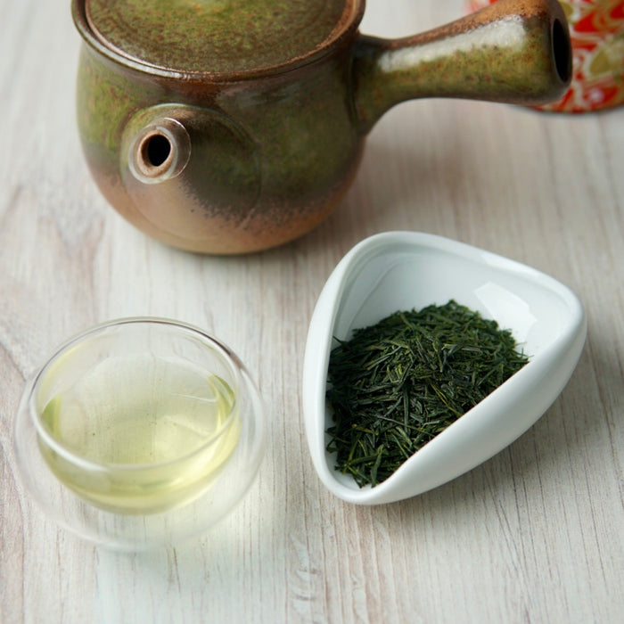 A ceramic green tea set on a light wood surface, featuring a small glazed teapot, a clear glass teacup with pale tea, and a white dish holding loose green tea leaves.