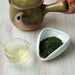 A ceramic green tea set on a light wood surface, featuring a small glazed teapot, a clear glass teacup with pale tea, and a white dish holding loose green tea leaves.