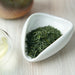 Green loose-leaf tea leaves displayed in a small white triangular ceramic dish on a light wood surface, with a blurred glass and ceramic vessel in the background.