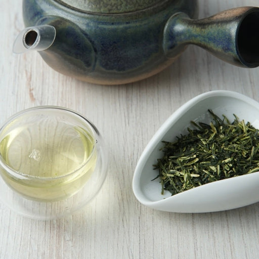 A ceramic green tea set on a light wood surface, featuring a dark blue-green teapot, a clear glass cup filled with pale tea, and a white dish holding loose green tea leaves.