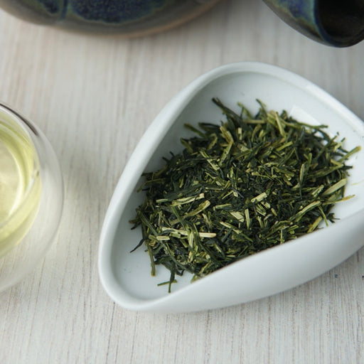 Dried loose-leaf green tea displayed in a small white dish on a light wood surface, with part of a glass cup and ceramic vessel visible in the background.