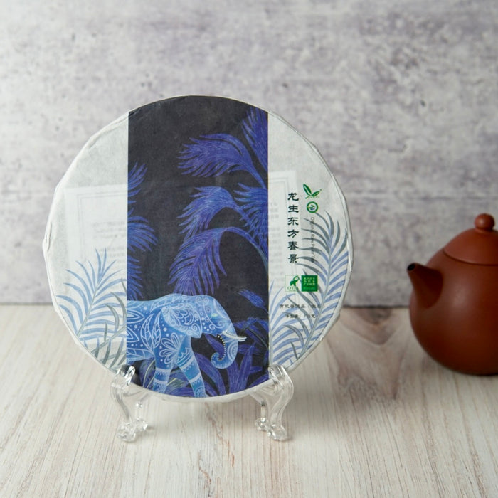 A round tea cake wrapped in decorative paper with a blue elephant and palm leaf design, displayed on clear stands on a light wood surface with a brown teapot in the background.