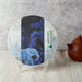 A round tea cake wrapped in decorative paper with a blue elephant and palm leaf design, displayed on clear stands on a light wood surface with a brown teapot in the background.