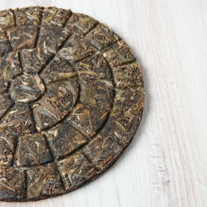 A close-up of a round compressed tea cake with visible leaf texture, placed on a light wood surface.