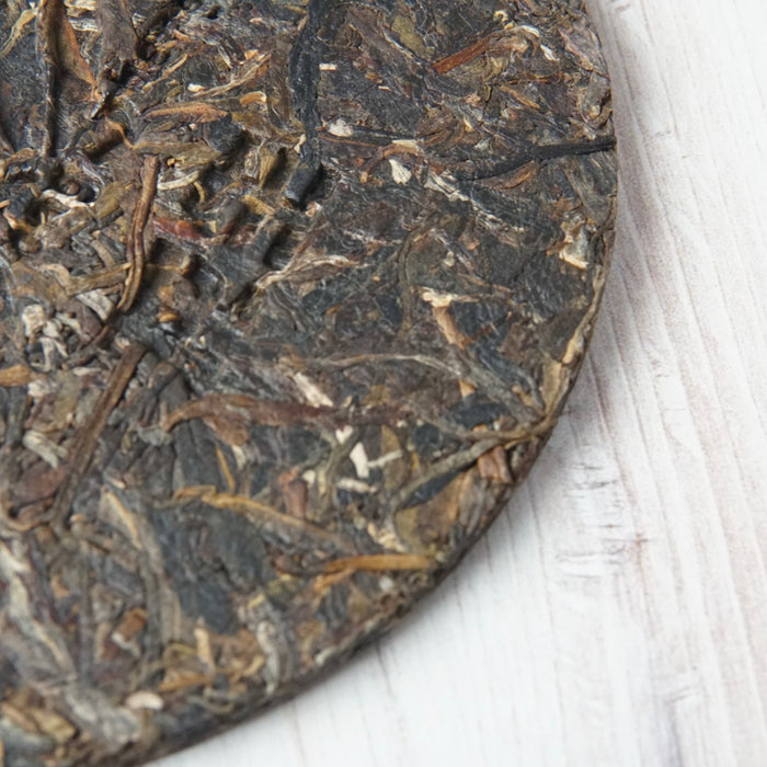 A close-up of a round compressed tea cake, showing the textured dark brown and black leafy surface, resting on a white painted wood background.