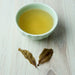 A small pale green ceramic cup filled with light yellow tea, placed on a light wood surface with two dried tea leaves in front.