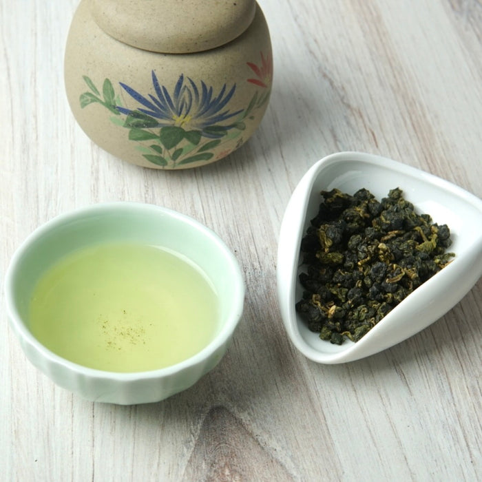 A small white ceramic bowl filled with pale yellow-green tea, a white dish holding dark rolled tea leaves, and a beige decorative ceramic jar in the background on a light wood surface.
