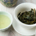 A white ceramic tea bowl filled with loose-leaf green tea leaves on a matching saucer, next to a small cup of pale brewed tea, on a light wood surface.