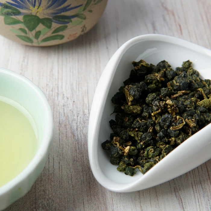 Rolled dark green tea leaves in a white ceramic dish, with a small cup of pale tea and part of a floral teacup on a light wood surface.