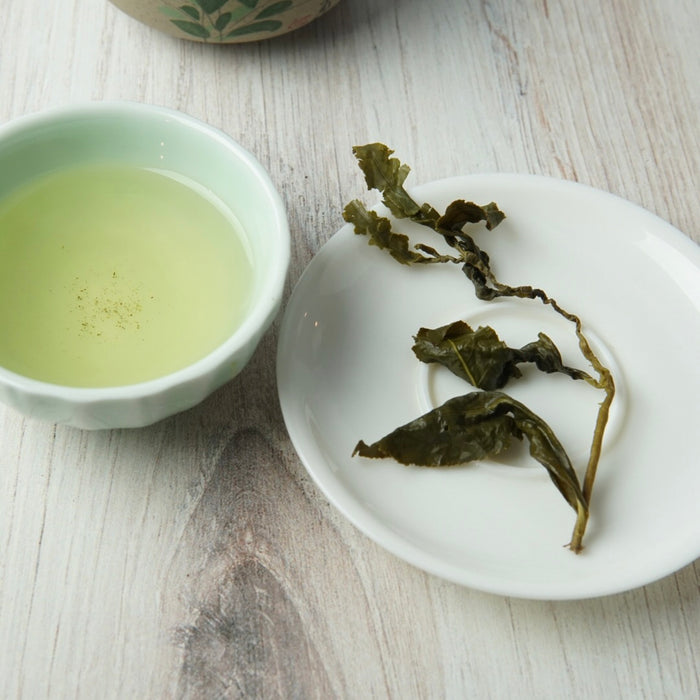 A small white ceramic bowl filled with pale green tea sits beside a white plate holding dried green tea leaves on a light wood surface.